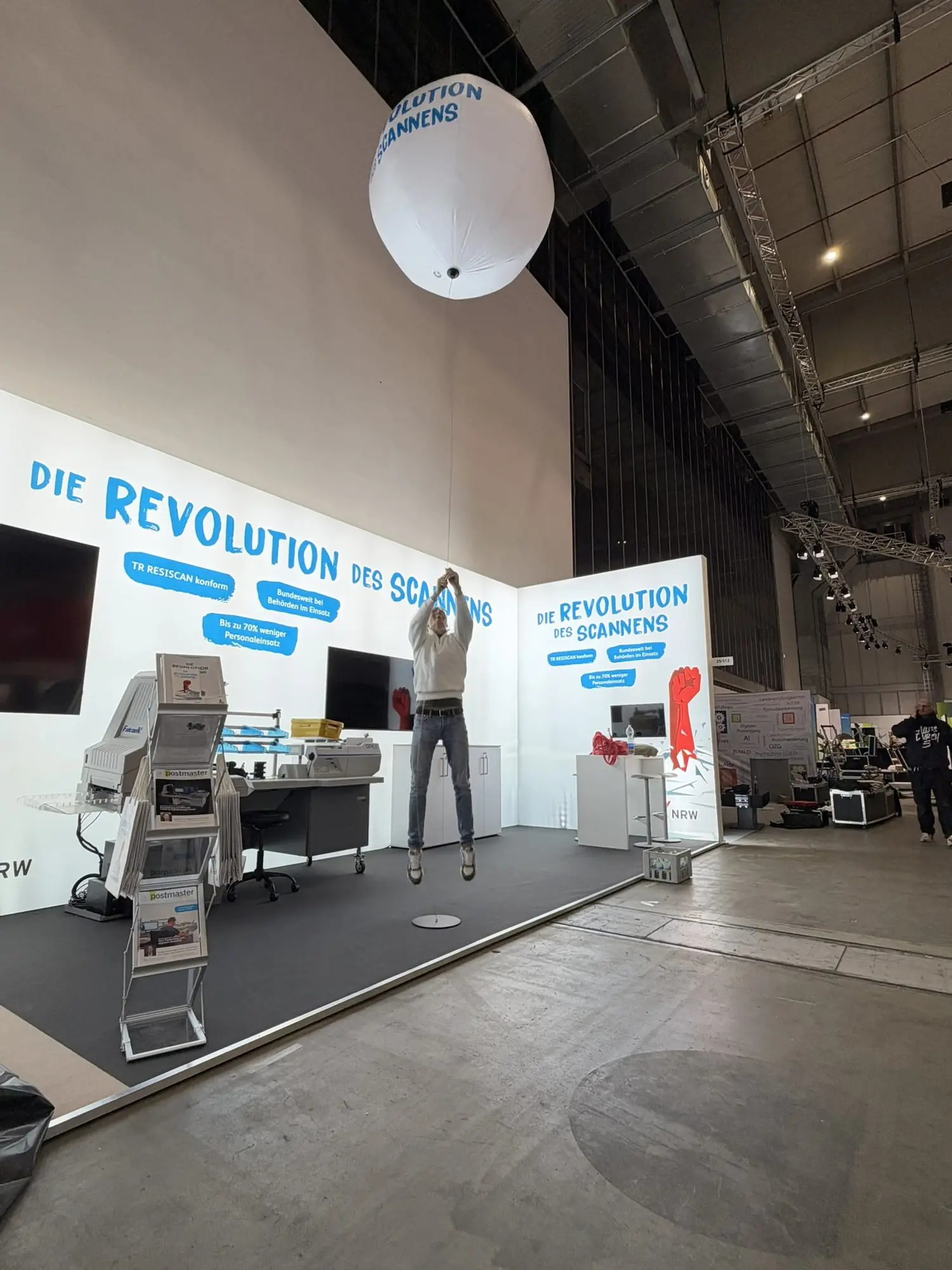 Fertiger X-NRW-Stand in Halle 25 mit OPEX-Scanarbeitsplatz, ein Mann springt am Seil des weißen Ballons mit dem Schriftzug Die Revolution des Scannens